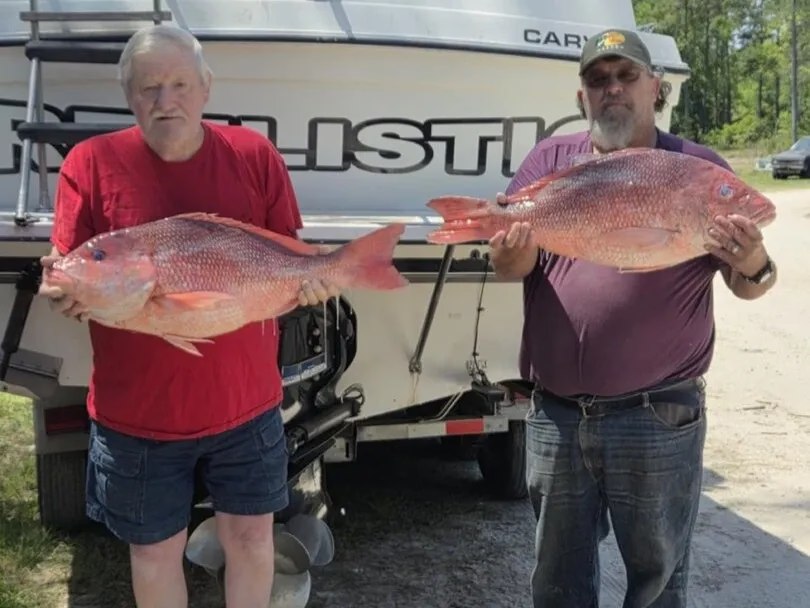 Two anglers displaying red fish beside a trailered boat
