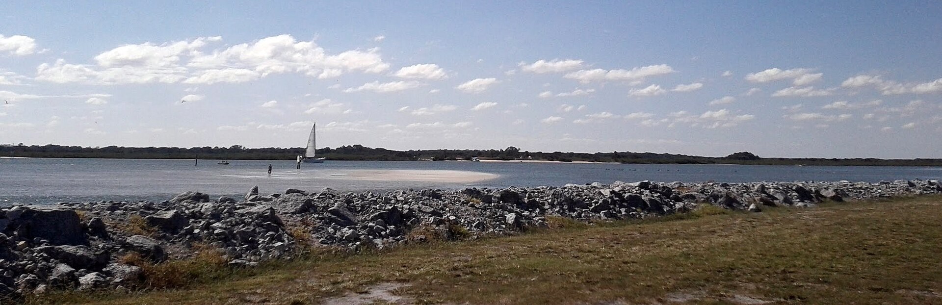 Rock-lined Ponce Inlet shoreline with a sailboat viewed from Lighthouse Point Park